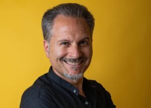 Smiling middle-aged man with gray hair and a goatee in a black shirt against a yellow background.